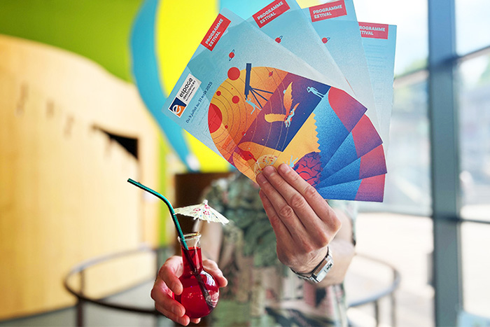 personne tendant devant lui les flyer de l'été et un ballon rempli d'eau coloré en rouge